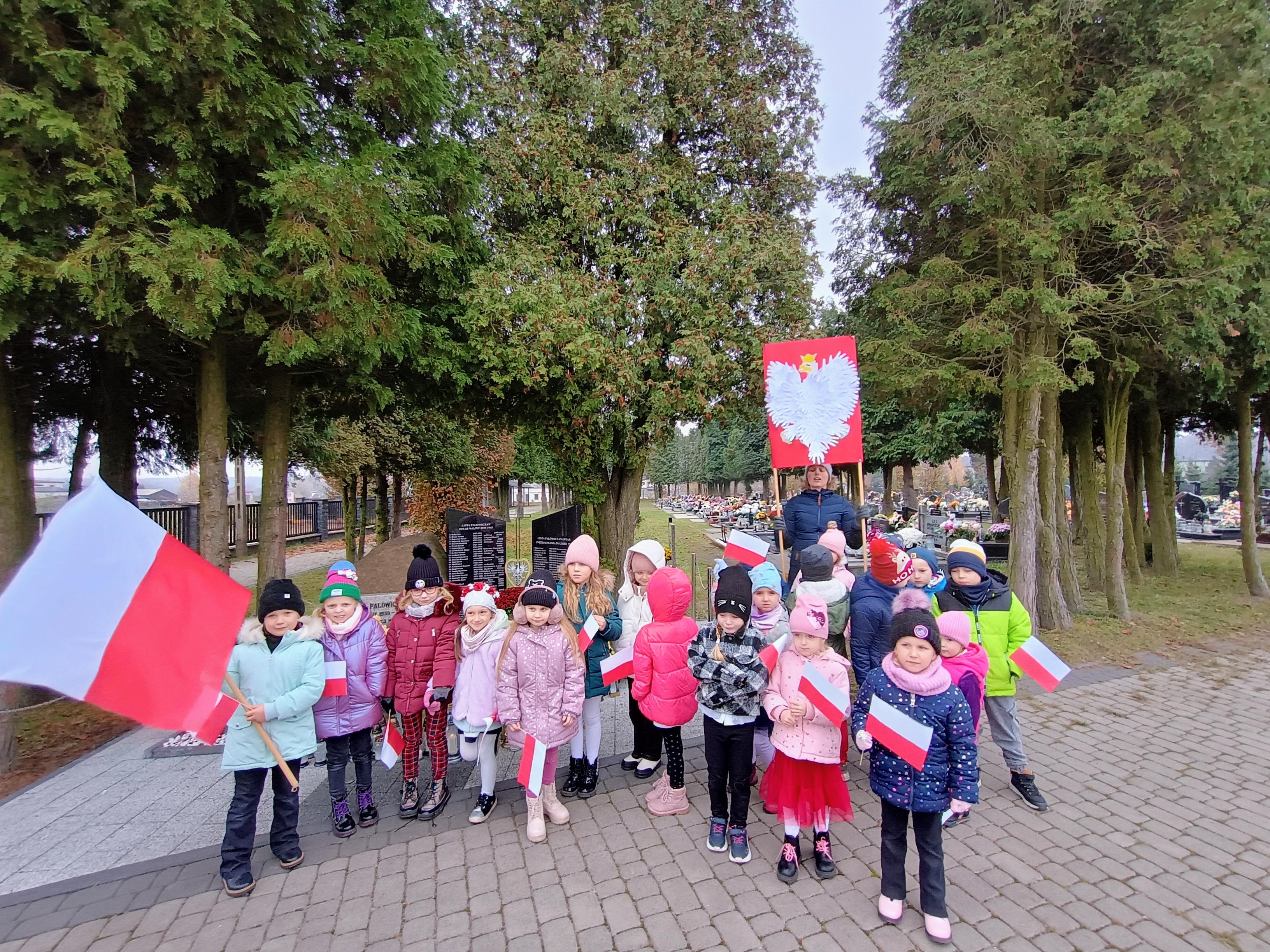 Grupa starszaków na tle pomnika ofiar. Trzymają w rękach flagi i wielkie godło Polski
